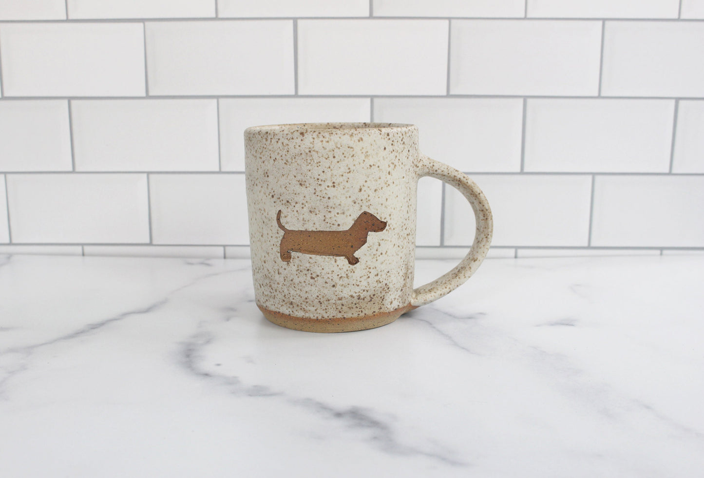 Speckled ceramic mug with dachshund silhouette on a marble surface with white tiled wall background