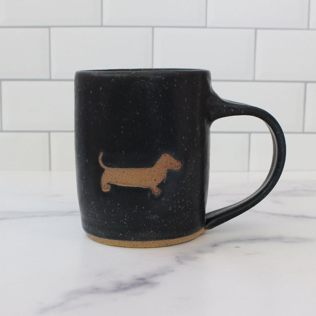 Black mug with a brown dachshund silhouette on a marble surface with a white tiled wall background